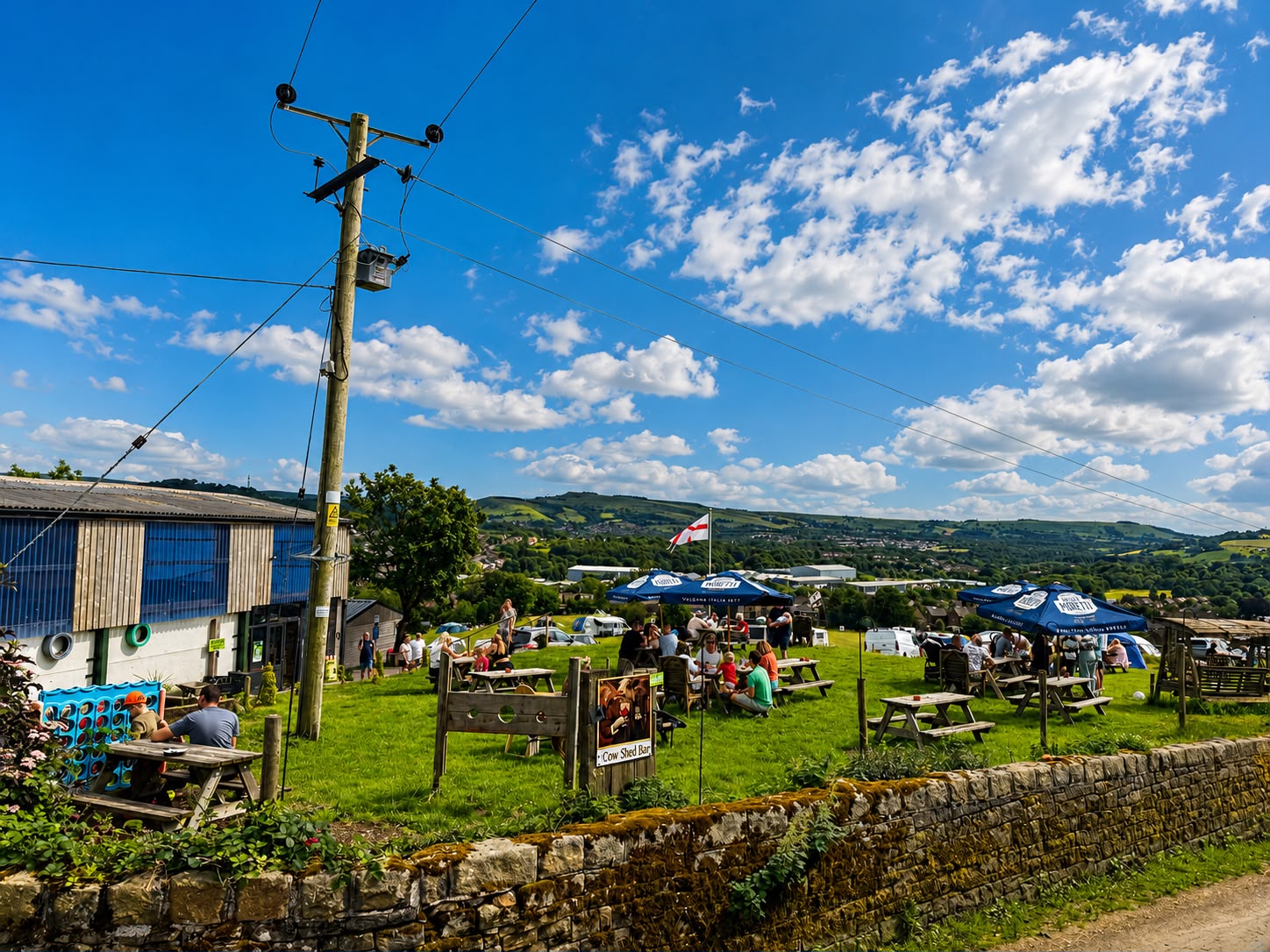 People relaxing at picnic benches and parasols outside The Cow Shed bar with the valley beyond